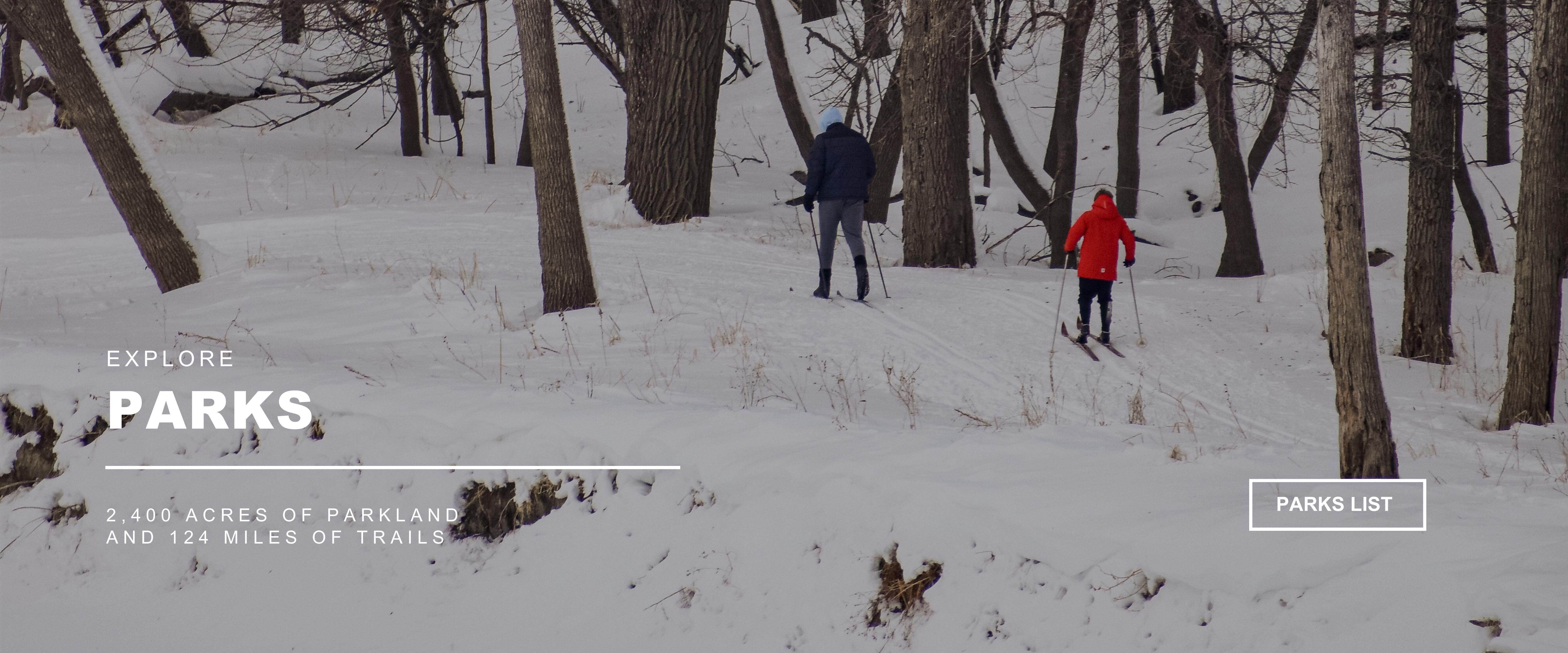 Explore PARKS - 2,400 acres of parkland and 124 miles of trails  - image of two people cross country skiing - one in a red winter coat the other in a navy blue coat with many trunks of trees in the background and snow on the ground - PARK LIST button