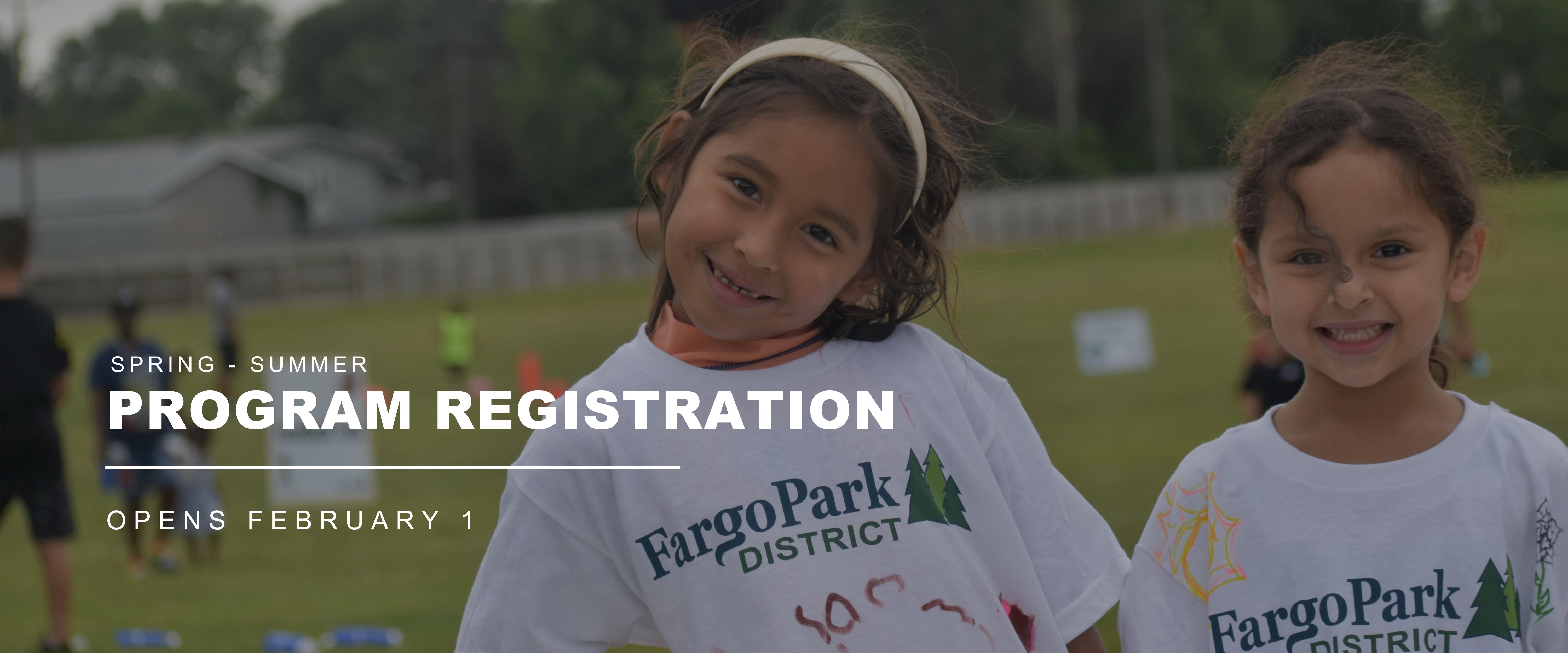 Spring-Summer Program Registration - Opens February 1 | Image of two young girls smiling wearing a Fargo Park District logoed white shirt, standing in front of kids playing soccer in the background