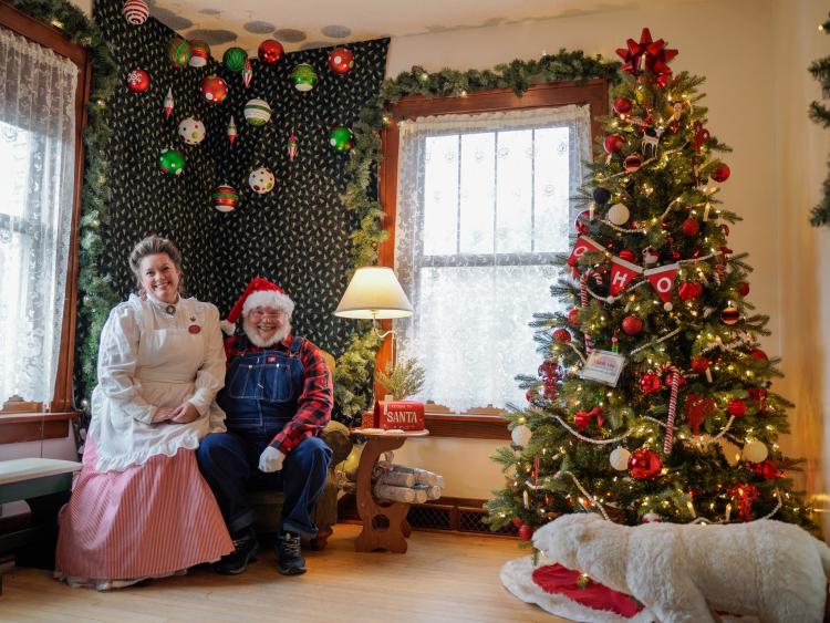 Santa and Mrs. Claus sitting in house on Santa's chair next to lamp and Christmas Tree decorated