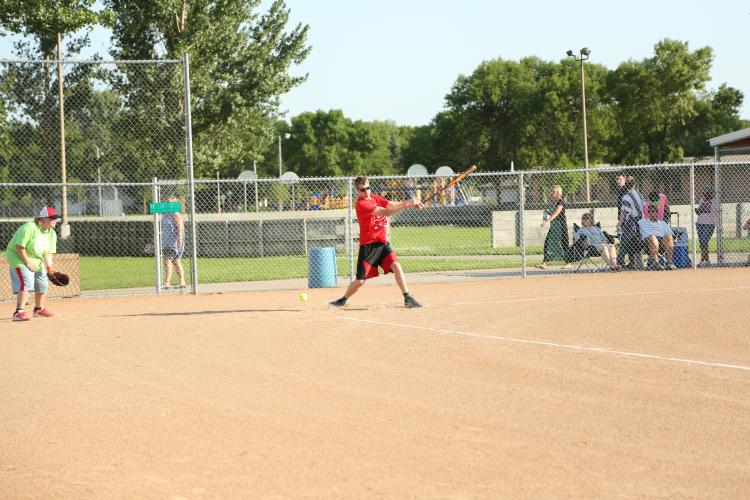 Male swinging softball bat on softball field with catcher standing behind.  Teammates and spectators in the background