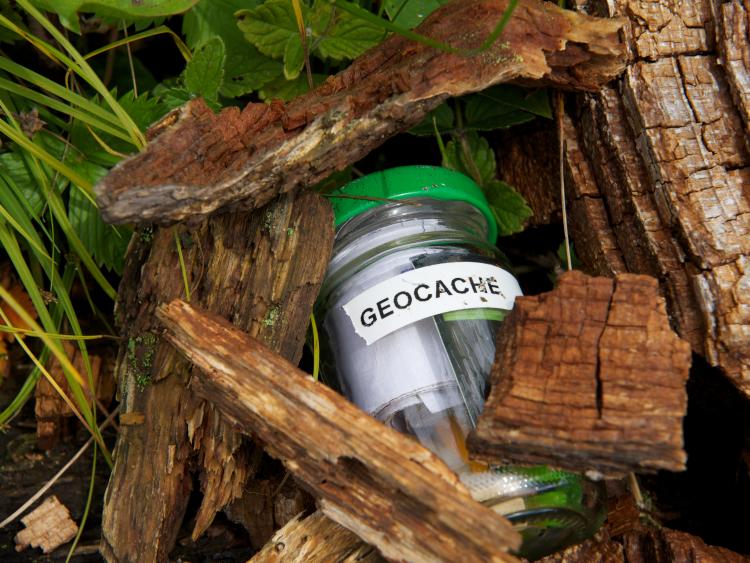 a jar with a green lid, jar labeled GEOCACHE with items in jar, semi-barried under wood