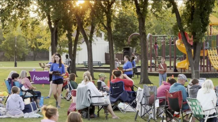 People sitting in lawn chairs and on blankets at Rheault Farm waiting for the Movie in the Park event to start.  Event Staff are walking around handing out clip boards for trivia.  The sun is setting in the background behind trees in the park
