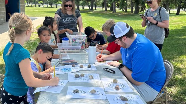 Man in blue Fargo Parks staff tshirt is sitting at a table helping manage the crafts projects happening while  6 children stand around the table working on the project and two adult females stand in the background watching