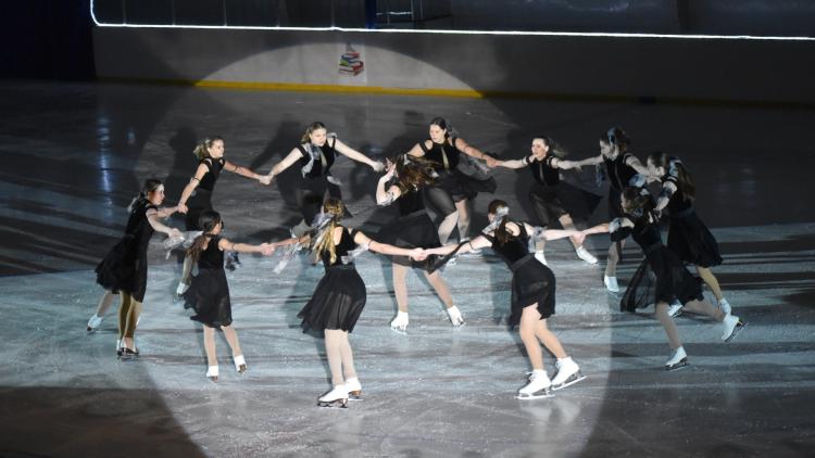 Ice Revue 11 older teens skating in a circle in black dresses around a girl in the middle skating in a spin, ice skating to a routine for the Fargo Parks Ice Revue
