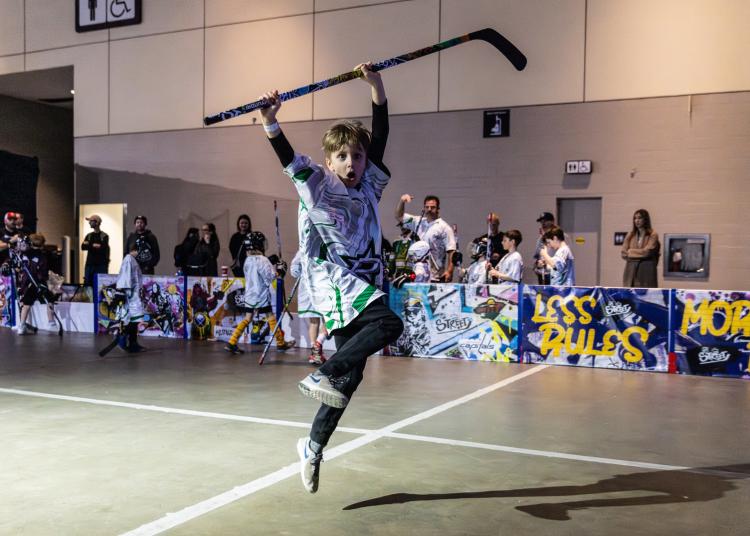 NHL STREET Child in jersey jumping in the air with a hockey stick over his head with parents and kids standing behind the boards cheering kids playing NHL Street on