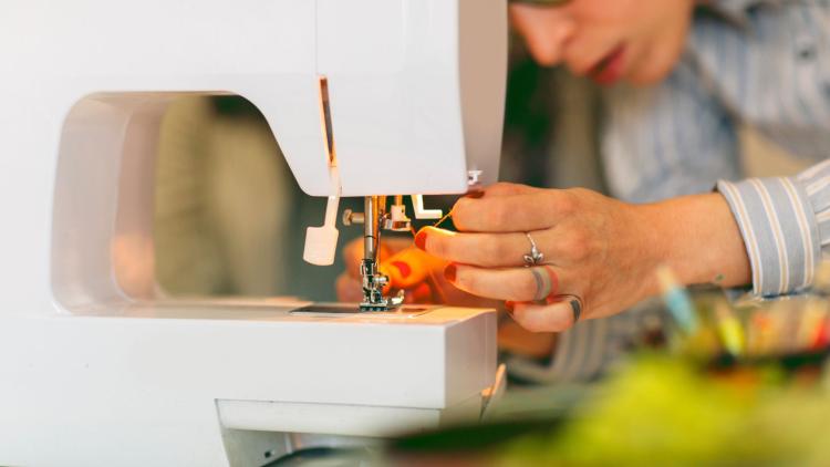 Close-up of a person threading a sewing machine needle, with their face partially visible in the background and sewing tools blurred in the foreground.
