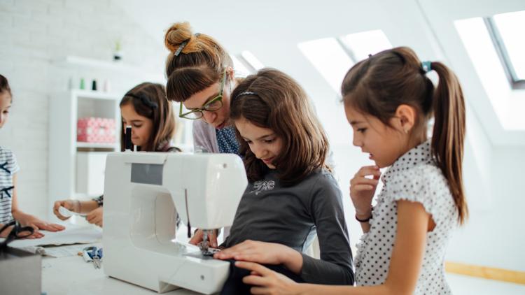Instructor guiding a group of young girls as they learn to use a sewing machine in a bright classroom.
