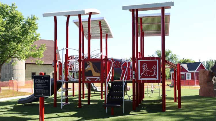 Rheault Farm Park .jpg Playground at Rheault Farm Park with a farm-themed design, featuring bright red structures topped with white roofs. Panels display images of a horse and a chicken, and a slide and climbing elements are visible. The playground sits on green turf with farm buildings and trees in the background on a sunny day.