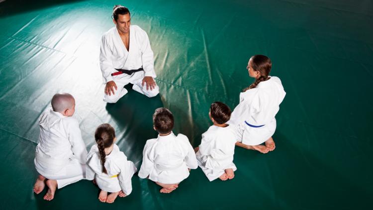 4 youth and instructor in Jiu Jitsu robes sitting on mat with their backs to camera in front of a male instructor