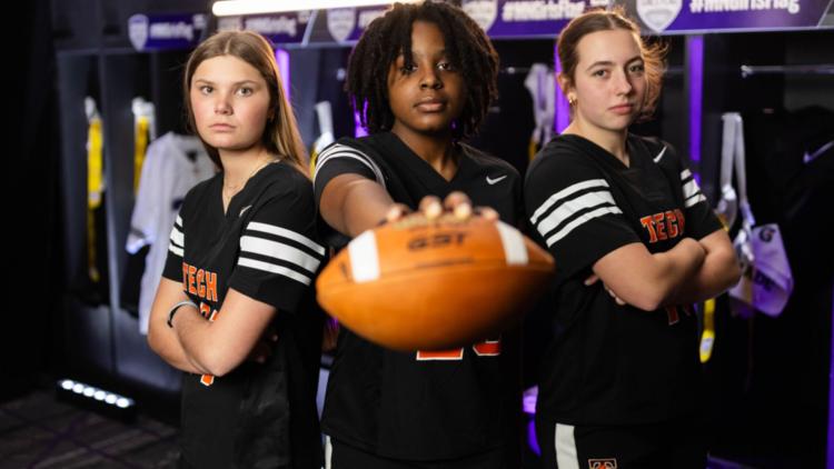Three high school girls in black football uniforms stand in a locker room, one holding a football toward the camera.