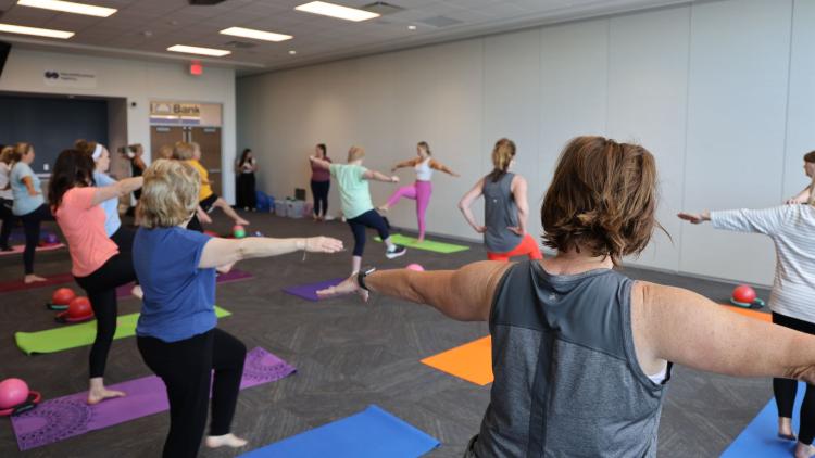 Older adults participate in a group fitness class, practicing balance and strength exercises on yoga mats in a community room.