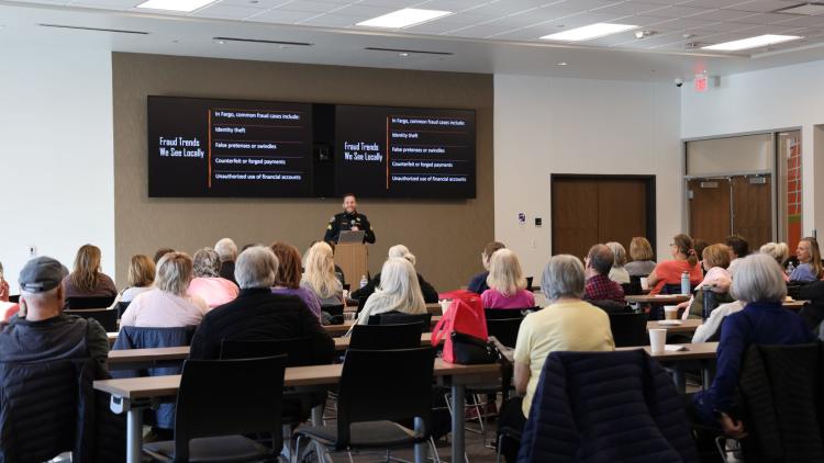 A police officer presents fraud prevention information to a group of older adults seated in a community meeting room.