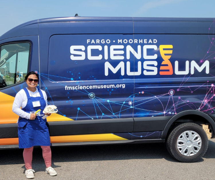 Woman standing in front of a large Fargo Moorhead Science Museum wrapped cargo van holding a beaver skull and magnifying glass