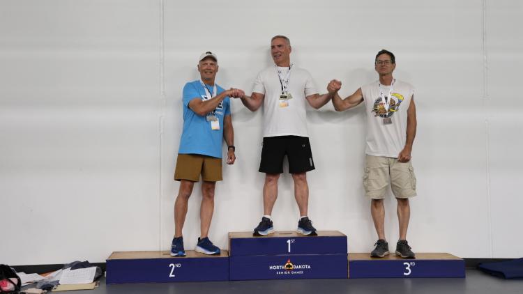 Three male athletes stand on a podium with medals for first, second, and third place at the North Dakota Senior Games.