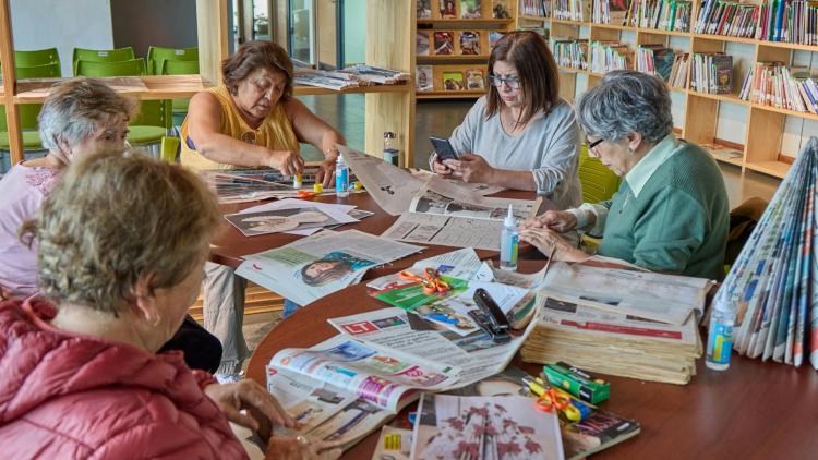 A group of older adults gathers around a table in a library for a craft activity, working with newspapers, scissors, glue and other materials.