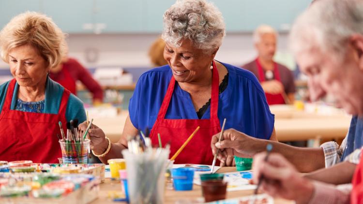 Older adults wearing red aprons participate in a group painting activity, using brushes and colorful paints around a shared table.