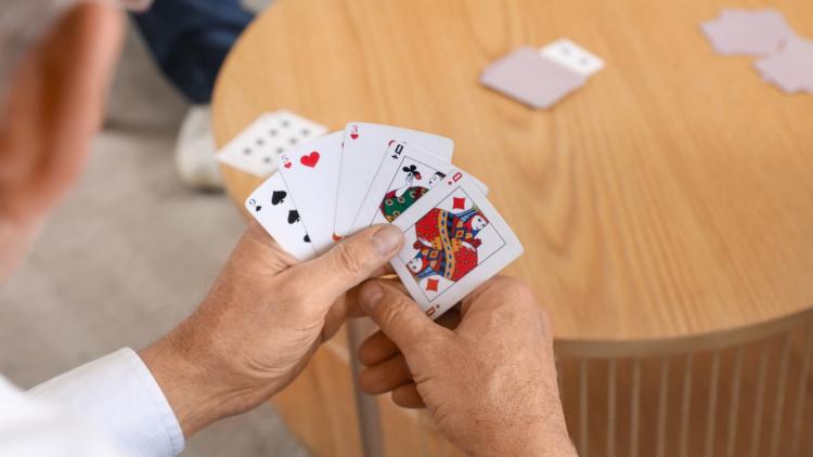 Close-up of a person holding a hand of playing cards over a round wooden table, with additional cards on the table in the background.