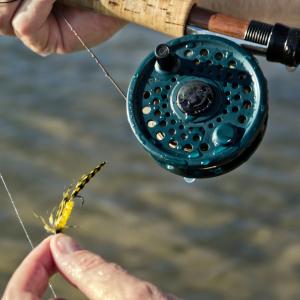 Close-up of hands holding a fly fishing rod and reel while tying a yellow-and-black fly lure, with water visible below.
