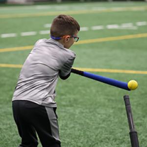 A child swings a bat to hit a ball off a tee during an adaptive sports activity on an indoor turf field.