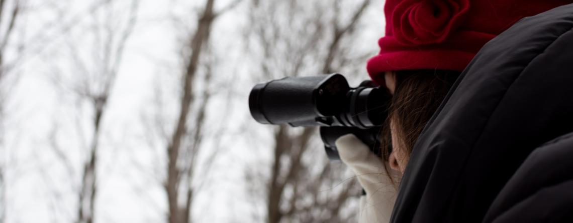 woman in red hat looking through binoculars in snowy forest