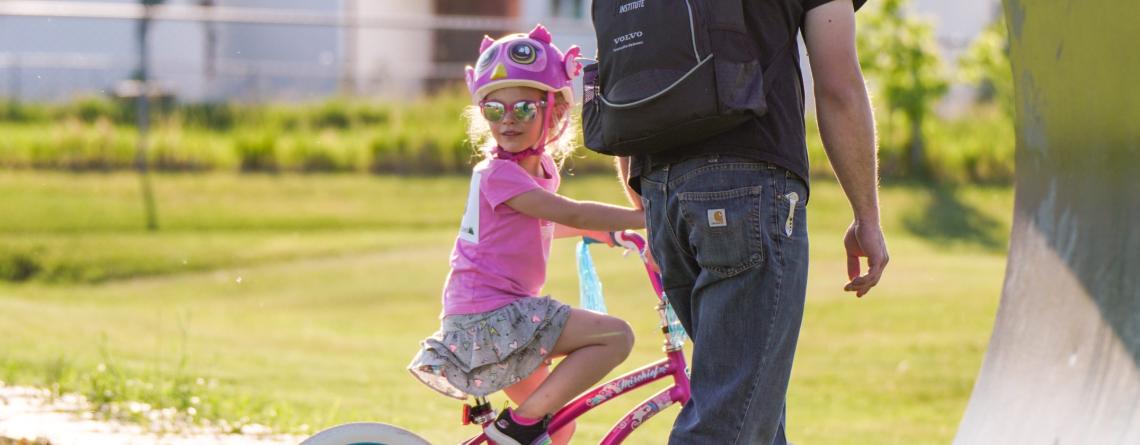 A young girl riding a bike at a park wearing a helmet with her dad walking next to her.