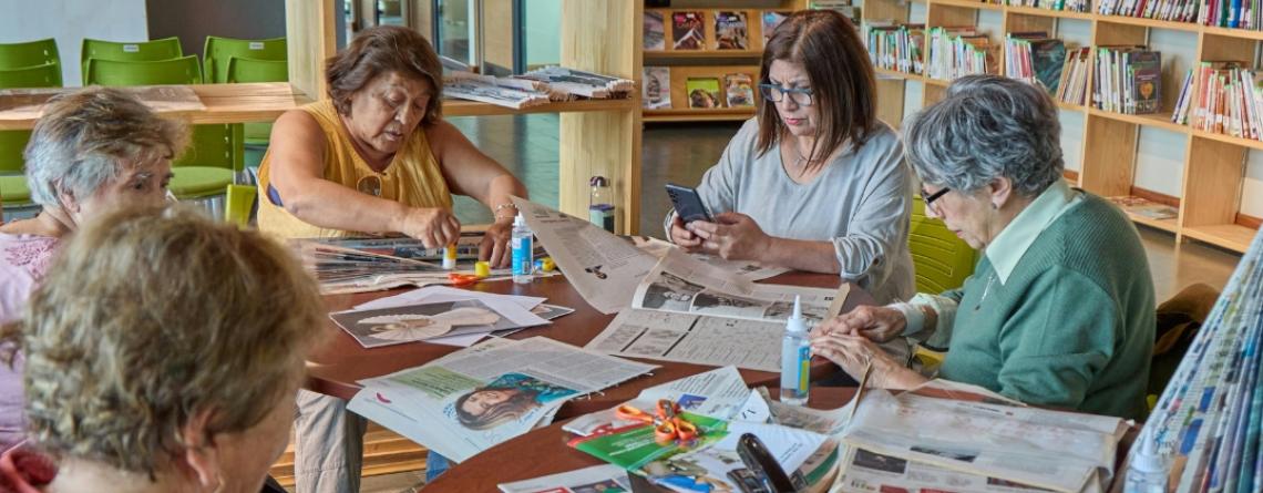 Group of older adults sitting around a table in a library or community center, working on crafts with newspapers, glue, and scissors, and chatting together.
