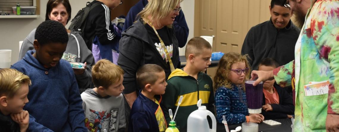 Group of children gathered around a science demonstration table, watching an instructor in a colorful lab coat show an experiment with a purple liquid, while adults observe in the background