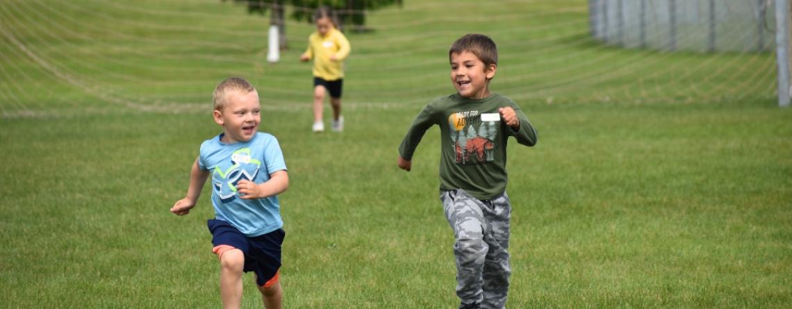 Two boys running through a grass field.  One little girl in the background running behind with a soccer net behind all.