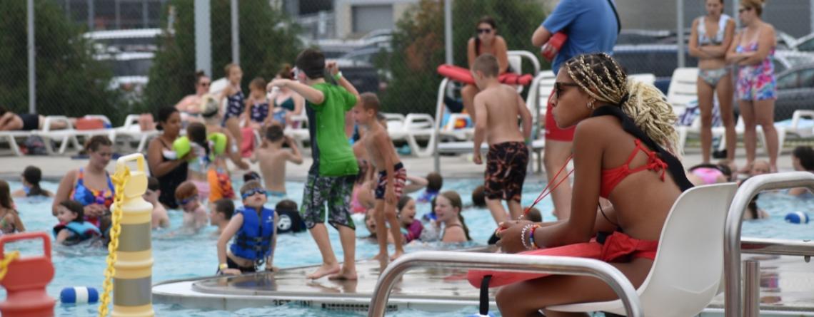 Children play and swim in a crowded outdoor pool while a lifeguard sits watching from a chair on the pool deck.