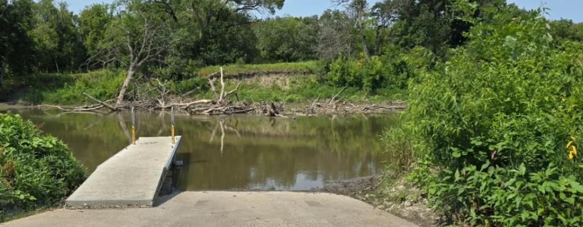 A concrete boat ramp extends into a calm river, surrounded by green trees and shrubs on a sunny summer day. Driftwood is visible along the opposite shoreline.