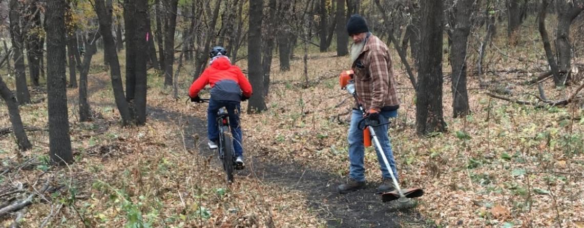 A cyclist rides along a narrow wooded trail while a park worker uses a string trimmer to maintain the trail. Fallen leaves cover the surrounding forest floor.