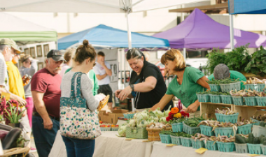 Photo shows two women assisting a woman shopping a vegetable booth at Red River Market. 