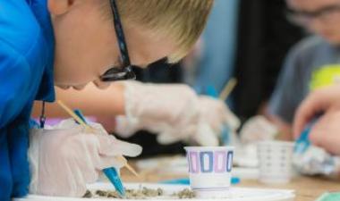 Child wearing glasses and gloves carefully works on a hands-on science project using tweezers and a tray of materials, with other children participating in the background.