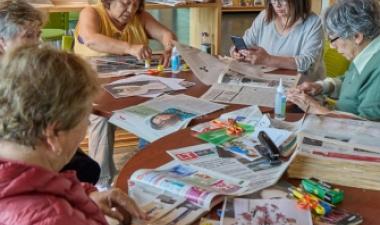 Group of older adults sitting around a table in a library or community center, working on crafts with newspapers, glue, and scissors, and chatting together.