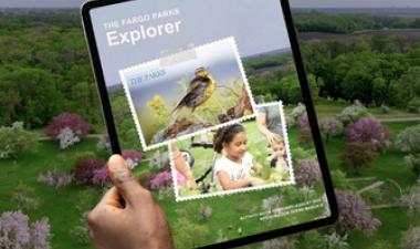 Person holding a tablet displaying The Fargo Parks Explorer activity guide, set against an aerial view of a green park landscape with trees and spring blossoms.uide, showing an aerial view of a park with green trees and flowering blossoms, alongside a person holding a tablet displaying the Explorer cover.