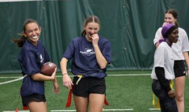 Four high school girls wearing flag football jerseys and belts walk and laugh together on an indoor turf field, one holding a football. All with mouthguards. 