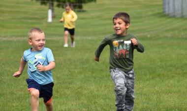 Two boys running through a grass field.  One little girl in the background running behind with a soccer net behind all.