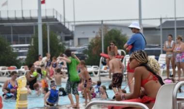 Children play and swim in a crowded outdoor pool while a lifeguard sits watching from a chair on the pool deck.