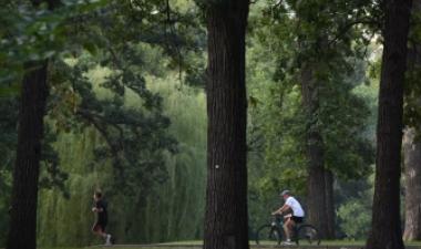 This image shows a person jogging and another person riding a bicycle along a paved trail through a wooded Fargo park.