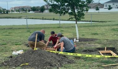 Three people work together on a landscaping project near a young tree, installing a wooden border around a planting area. A pile of soil, tools and caution tape surround the work area.