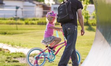 A young girl riding a bike at a park wearing a helmet with her dad walking next to her.