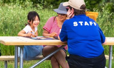 A young girl and her mother doing crafts at an outdoor picnic table in the park with a staff member. 