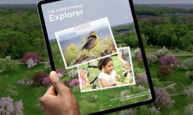 Person holding a tablet displaying The Fargo Parks Explorer activity guide, set against an aerial view of a green park landscape with trees and spring blossoms.uide, showing an aerial view of a park with green trees and flowering blossoms, alongside a person holding a tablet displaying the Explorer cover.