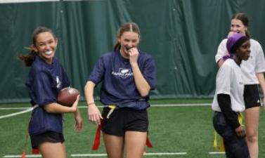 Four high school girls wearing flag football jerseys and belts walk and laugh together on an indoor turf field, one holding a football. All with mouthguards. 