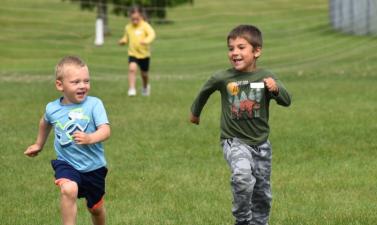 Two boys running through a grass field.  One little girl in the background running behind with a soccer net behind all.