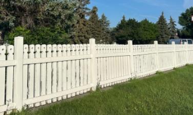 A long white picket fence stretches across a grassy area at Yunker Farm Park, with mature trees, a blue sky and a parked vehicle in the background.