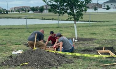 Three people work together on a landscaping project near a young tree, installing a wooden border around a planting area. A pile of soil, tools and caution tape surround the work area.
