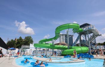 Outdoor water park on a sunny day with a large bright-green waterslide twisting above a lazy river. Many children and adults float in blue inner tubes or swim in the pool, while lifeguards stand nearby. A tall staircase leads to the slide entrance, and more people wait in line at the top. Trees, buildings, and a partly cloudy blue sky are visible in the background.