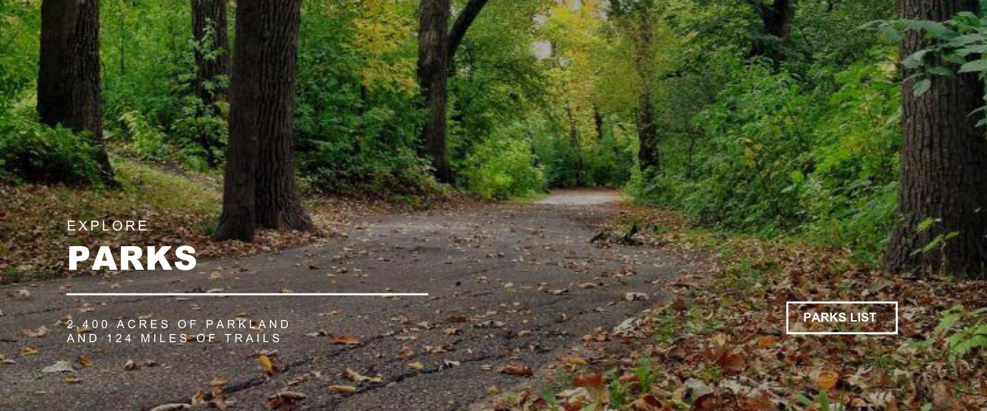 an asphalt surfaced trail going through trees that are green and turning yellow with fall leaves partially covering the surface of the trail, EXPLORE PARKS, PARKS LIST Button