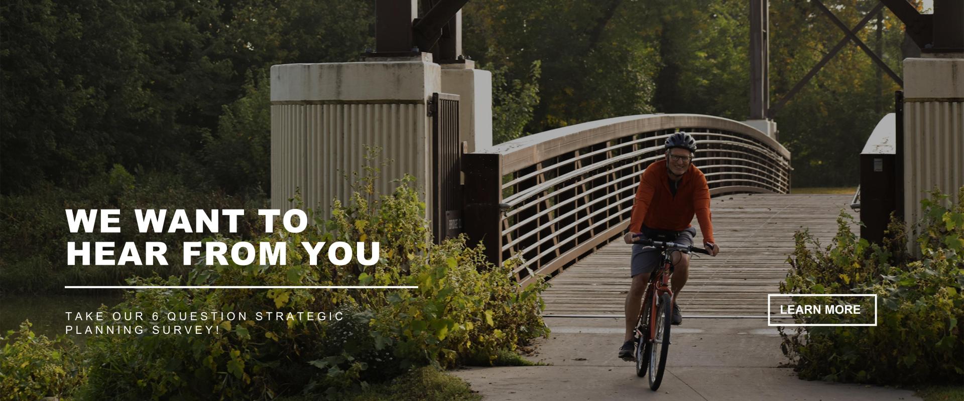 WE WANT TO HEAR FROM YOU - Take our 6 question strategic planning survey - Man on bike, traveling over the Lindenwood bridge with greenery and trees in the background